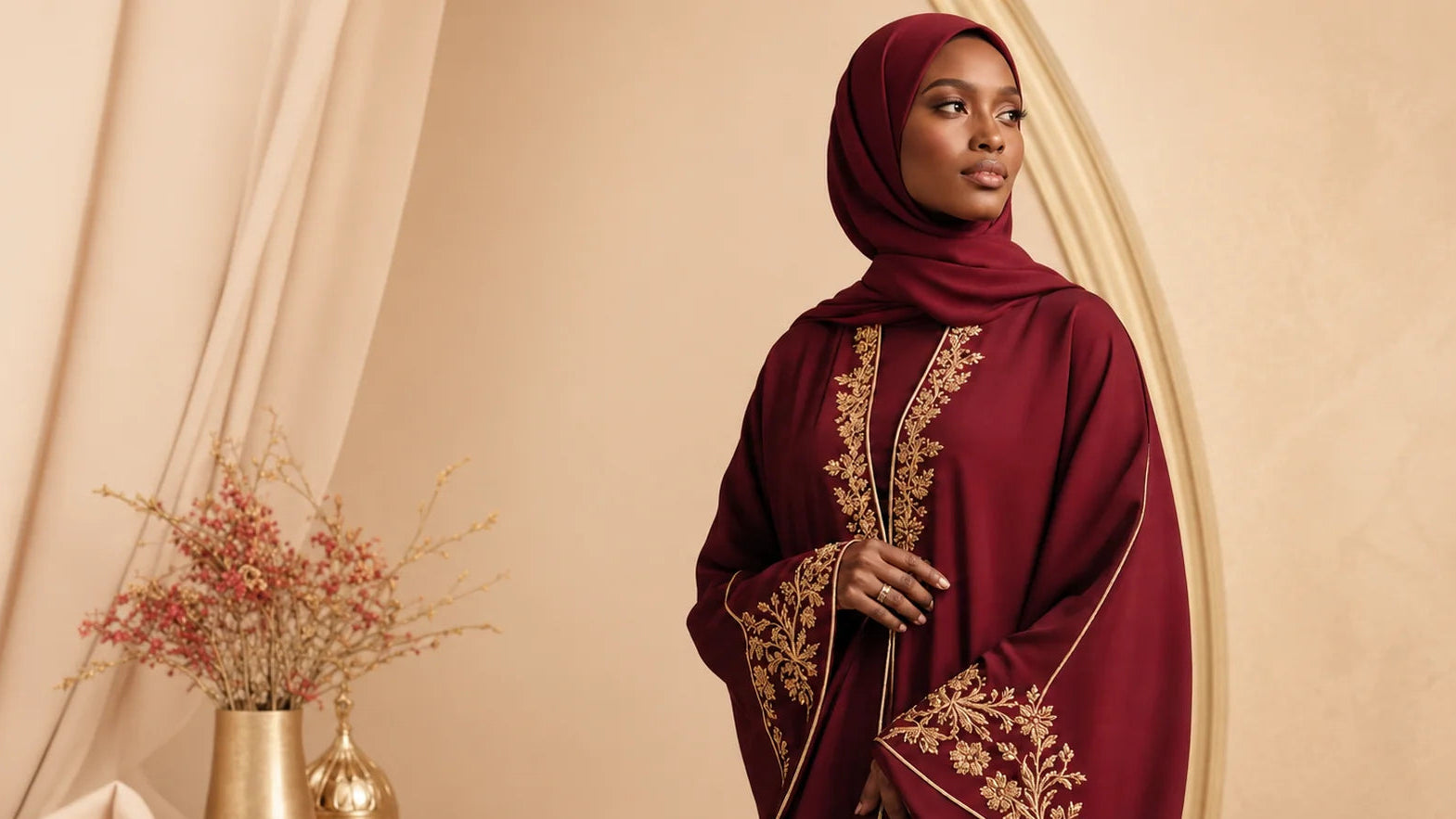 Woman wearing a burgundy embroidered hijab against a beige curtain background